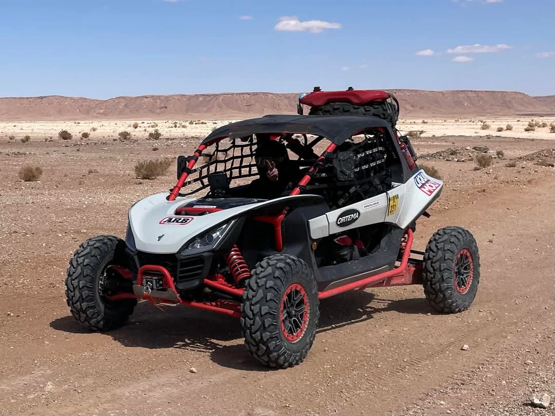 Ein weiß-roter Side-by-Side-Offroad-Buggy mit Sicherheitskäfig, parkend in einer trockenen, steinigen Wüstenlandschaft mit spärlicher Vegetation und Bergen im Hintergrund. Es befindet sich eine Person im Fahrzeug, die jedoch aufgrund des Käfigs nur schwer zu erkennen ist.