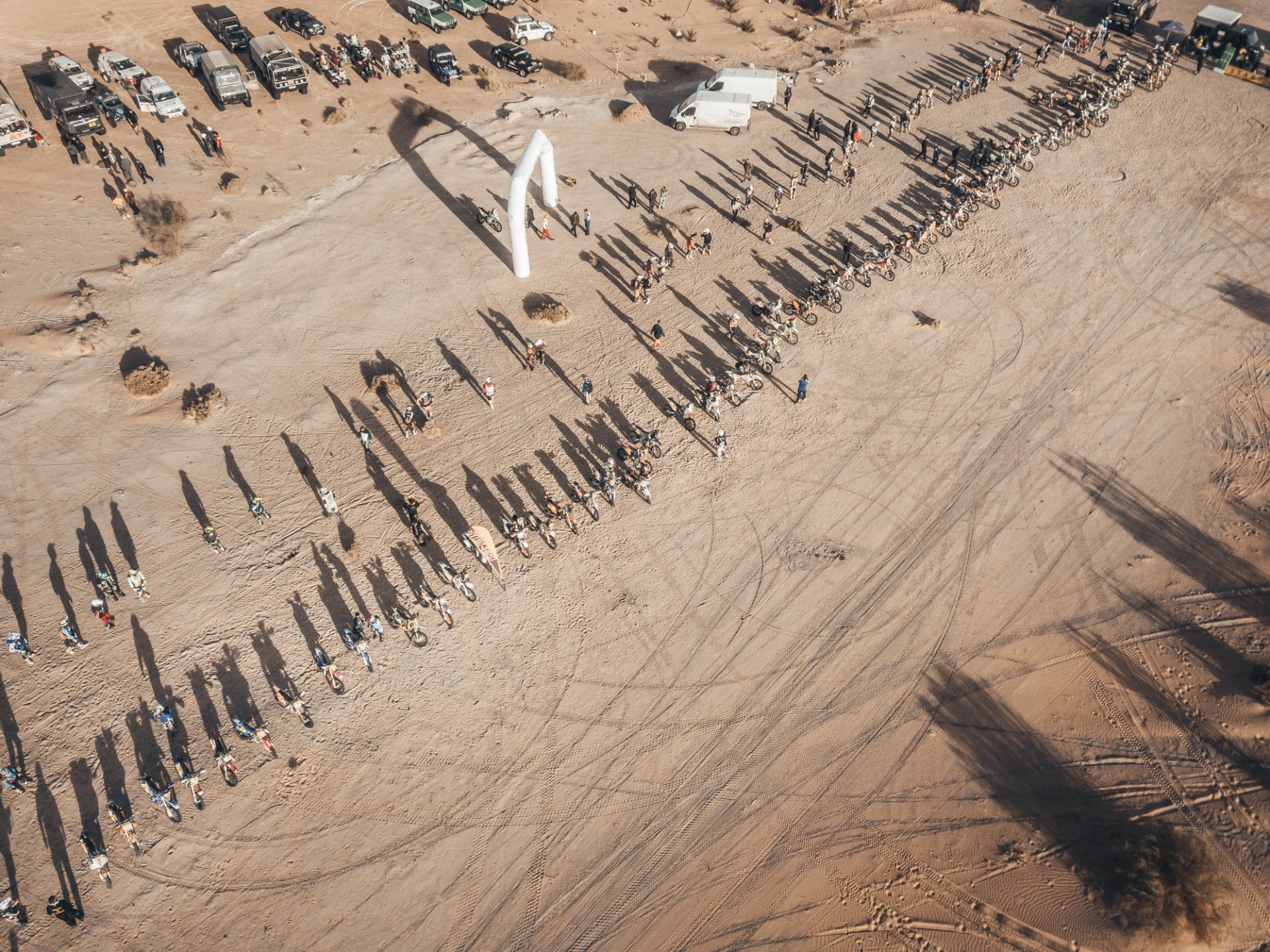 Luftaufnahme einer Wüstenlandschaft mit einer langen Schlange von Menschen, die im Sand verteilt stehen, und deren Schatten, die sich lange auf dem Boden abzeichnen, begleitet von einigen Fahrzeugen im Hintergrund. Ein weißes aufgeblasenes Bogen-Objekt am oberen Bildrand und mehrere parkende Autos deuten auf eine Veranstaltung oder Aktivität hin.
