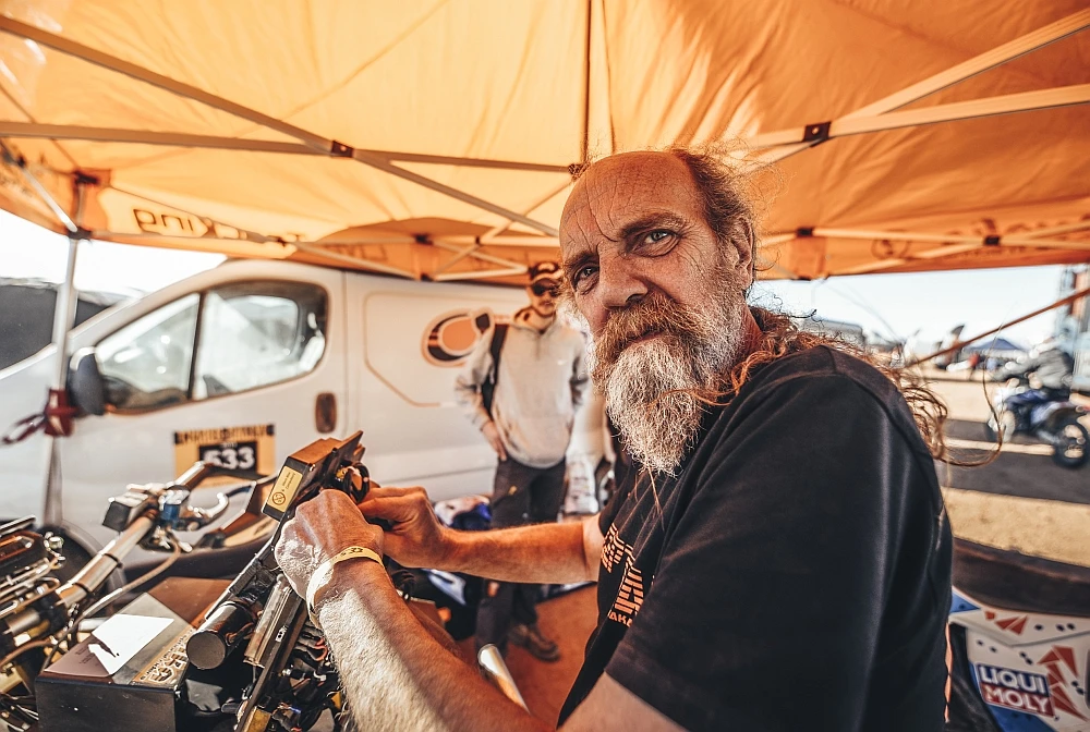 Tuareg Rallye Mechaniker Ein ernst dreinblickender Mann mit vollem Bart und langen Haaren arbeitet an einer Motorradkomponente unter einem orangefarbenen Zelt. Im Hintergrund sind weitere Menschen und ein weißer Van zu erkennen. Die Stimmung ist fokussiert und professionell in einer Motorsportumgebung.
