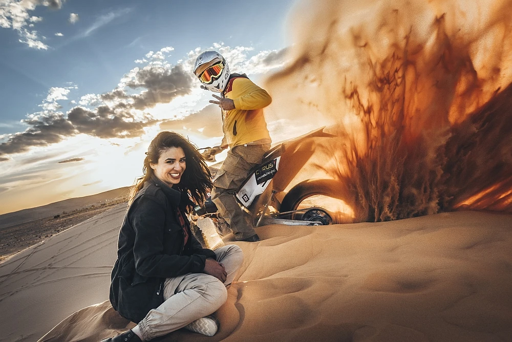 Tuareg Rallye Teilnehmer Eine Frau sitzt lächelnd auf einer Sanddüne, während ein Mann in Motorradkleidung neben einem Offroad-Motorrad steht. Er erzeugt eine Sandwolke mit dem Hinterreifen, was eine dynamische und abenteuerliche Atmosphäre in der Wüstenlandschaft unter einem beeindruckenden Wolkenhimmel schafft.