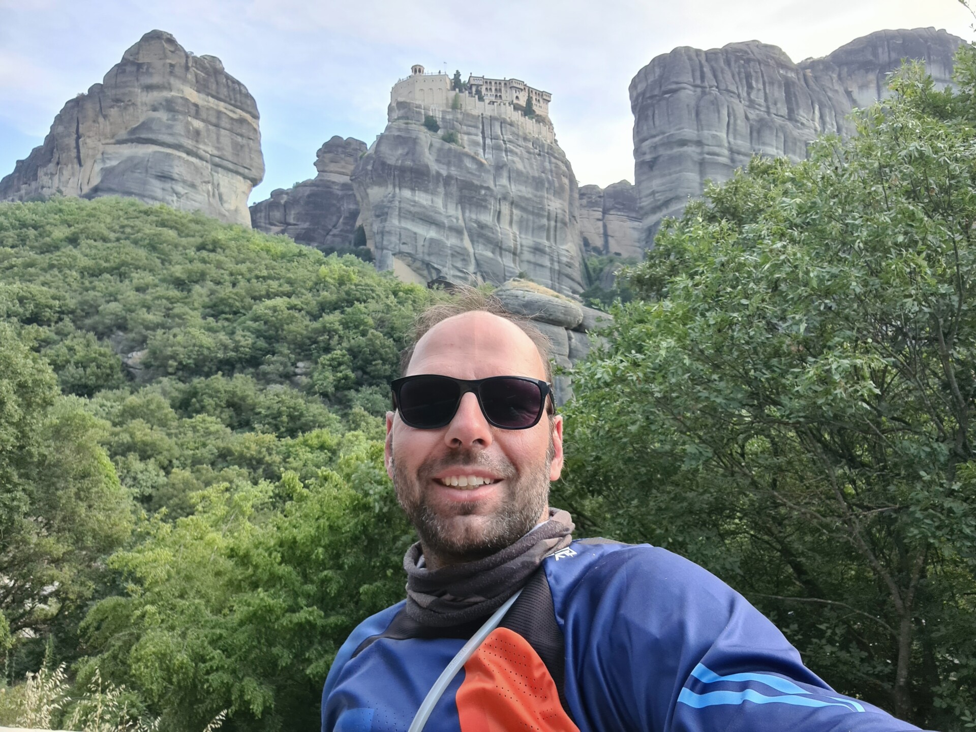 Ein Mann mit Sonnenbrille und Rucksack nimmt ein Selfie vor den imposanten Felsformationen von Meteora, Griechenland, mit einem dichten Wald im Vordergrund.