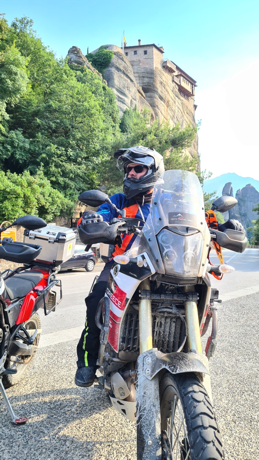 Ein Motorradfahrer sitzt auf einem voll bepackten Reisemotorrad vor der beeindruckenden Kulisse eines auf einer hohen Felsformation errichteten Klosters. Der Motorradhelm ist geöffnet und der Fahrer schaut in die Kamera, während im Hintergrund das grüne Laub und die historischen Mauern des Klosters sichtbar sind.