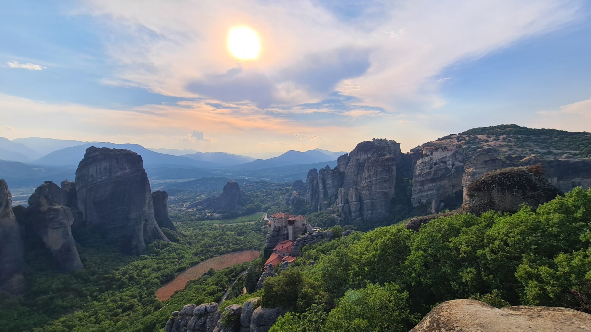 Blick auf die beeindruckenden Felsformationen und Klöster von Meteora in Griechenland bei Sonnenuntergang, mit sichtbarem üppigem Grün und hügeligen Landschaften im Hintergrund.