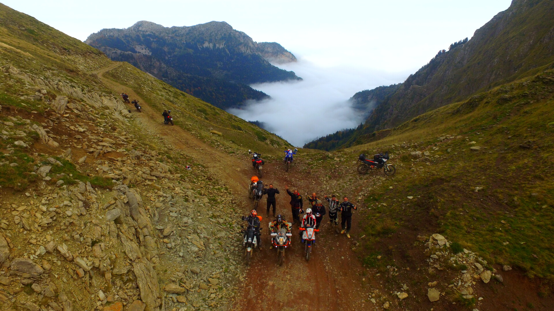 Eine Gruppe von Motorradfahrern macht eine Pause auf einem Bergpfad. Sie stehen oder sitzen neben ihren Motorrädern und genießen die Aussicht auf ein tiefes Tal, das von dichtem Nebel bedeckt ist. Die umliegenden Berghänge sind karg und steinig.