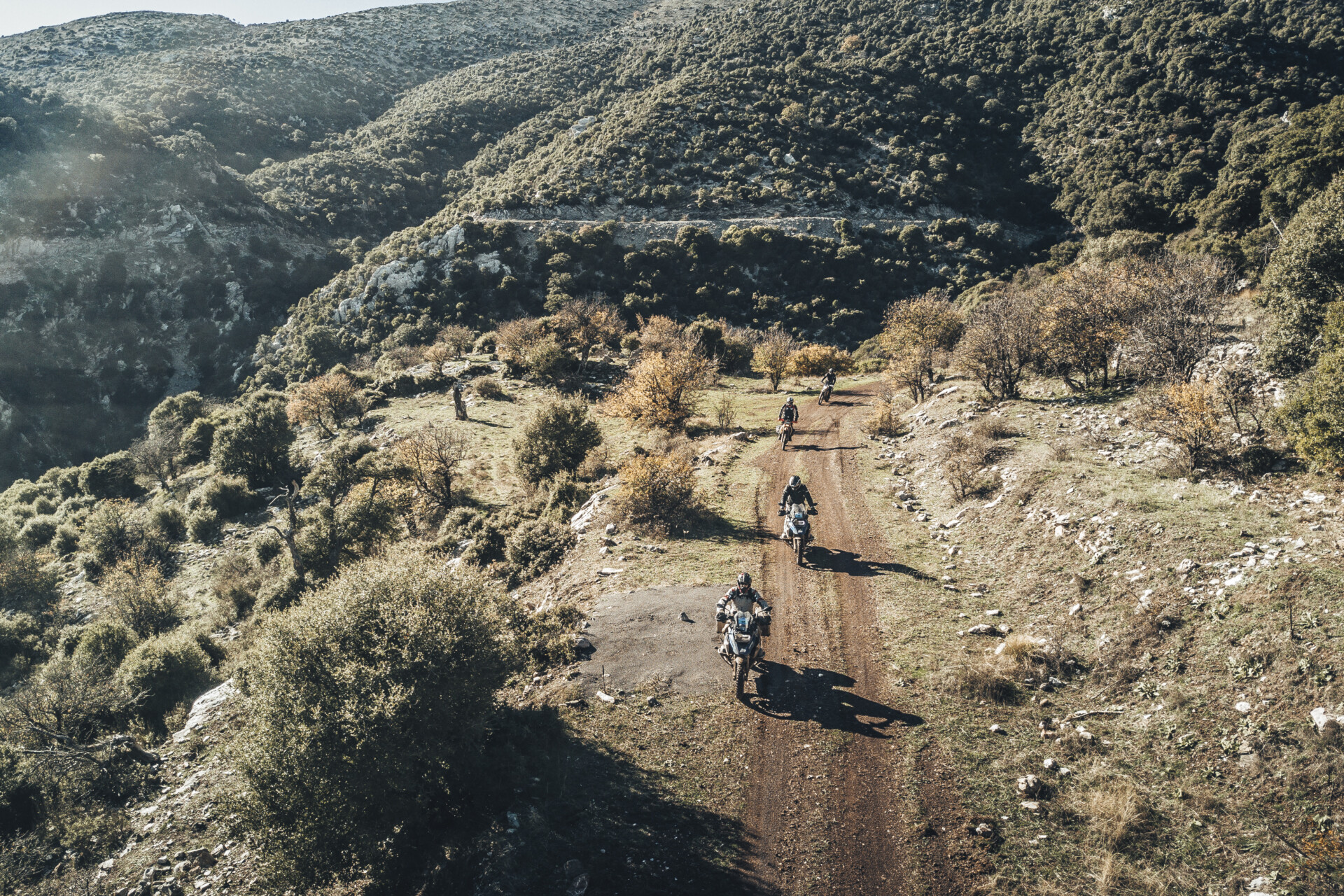 Luftaufnahme einer Gruppe von Motorradfahrern, die auf einer Schotterstraße in einer hügeligen, bewaldeten Landschaft fahren. Die Szene zeigt das ländliche Gebiet bei sonnigem Wetter, wobei das Licht die Textur des Geländes und der Vegetation hervorhebt.