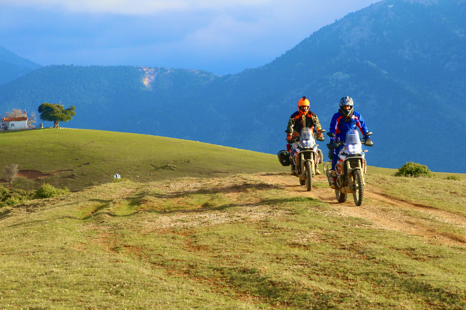 Biker im Gebirge Zwei Motorradfahrer auf Geländemotorrädern fahren auf einem Feldweg in einer hügeligen Landschaft. Sie tragen volle Schutzausrüstung, darunter Helme und Jacken. Im Hintergrund sind bewaldete Berge und eine kleine Kapelle auf einem grünen Hügel zu sehen. Die Szene wird von einem klaren Himmel mit einigen Wolken beleuchtet.