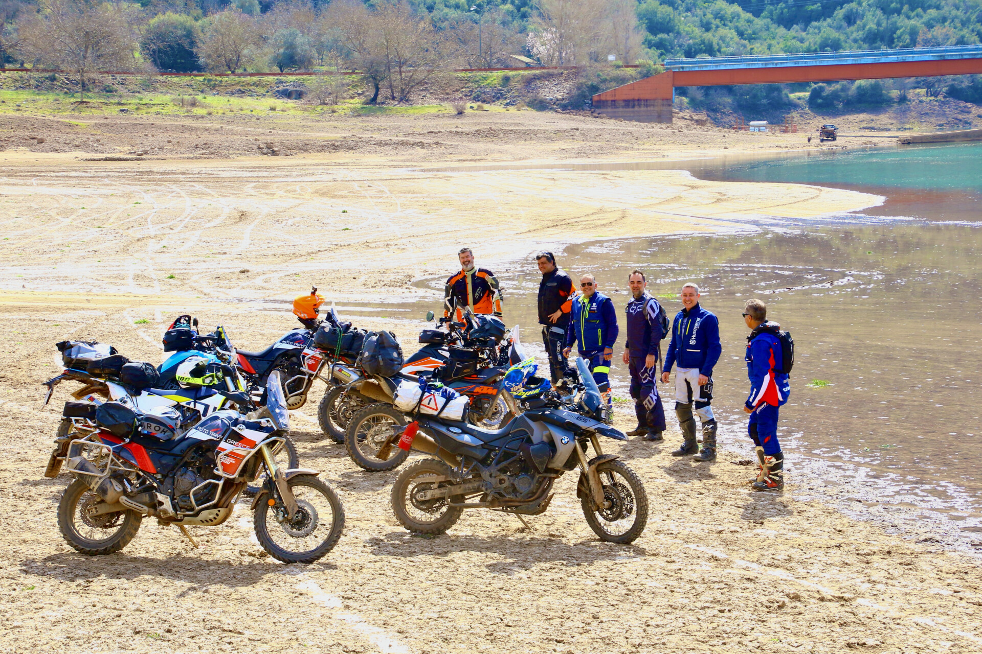 Gruppe von Motorradfahrern pausiert an einem sandigen Ufer mit ihren Touring-Motorrädern im Vordergrund, im Hintergrund ist ein ruhiger See und eine Brücke sichtbar.