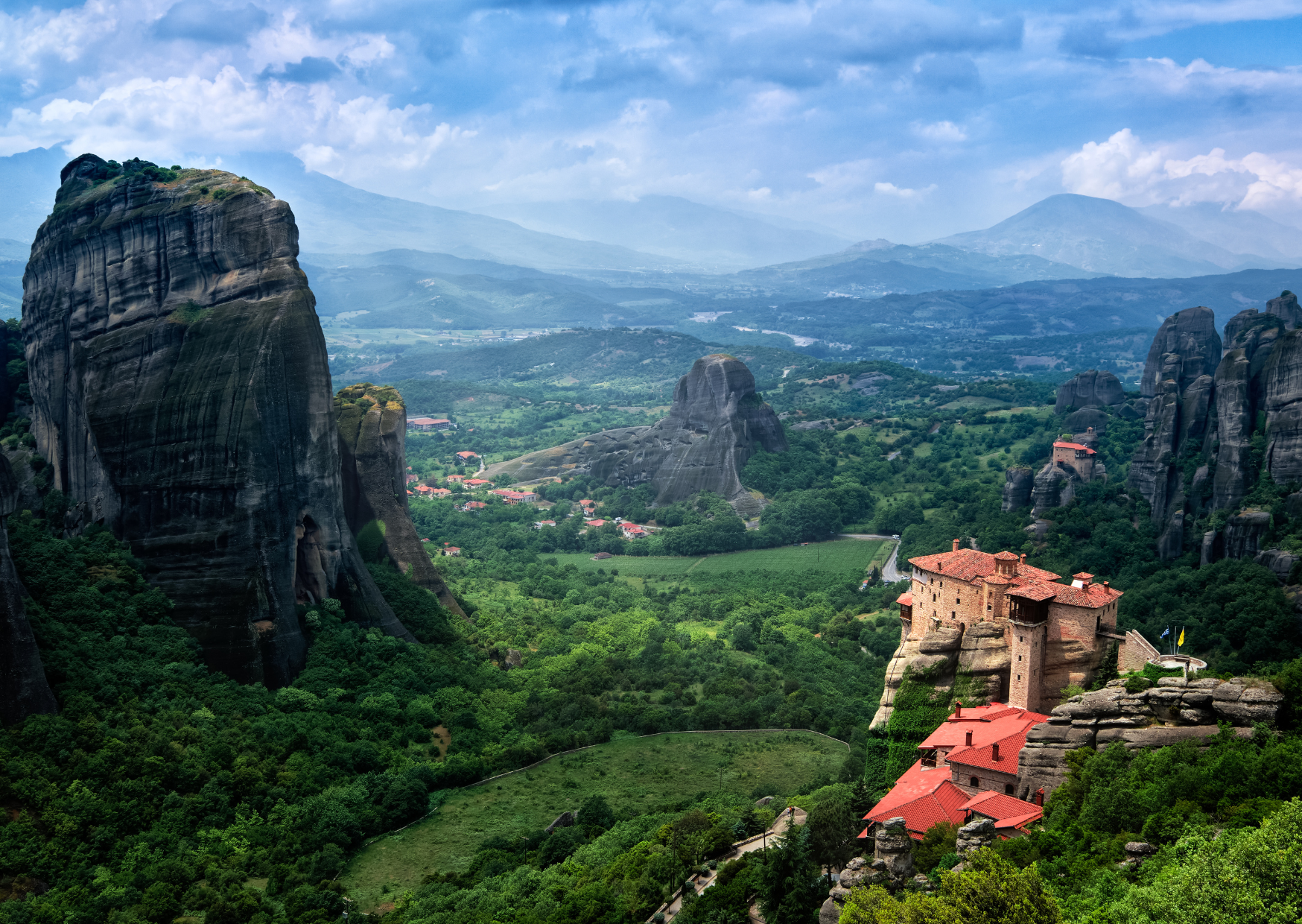 Panorama der Meteora-Klöster in Griechenland, gezeigt an einem bewölkten Tag. Markante, steil aufragende Felsformationen umgeben von üppigem grünem Wald, mit einem traditionellen Kloster, das prominent auf einer dieser Felsen sitzt. Im Hintergrund verschwimmen weitere Hügel und Berge in den Dunst des Horizonts.