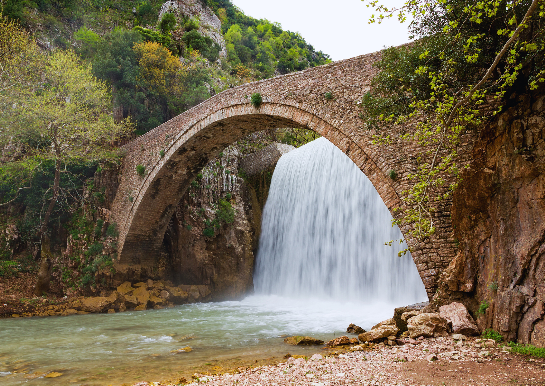 Bild eines alten Steinbogens über einem Fluss, durch den Wasser sprudelt und einen Wasserfall bildet. Das Bild zeigt sowohl das klare Wasser des Flusses als auch die bewachsenen Felswände und die grüne Vegetation, die den Ort umgibt.